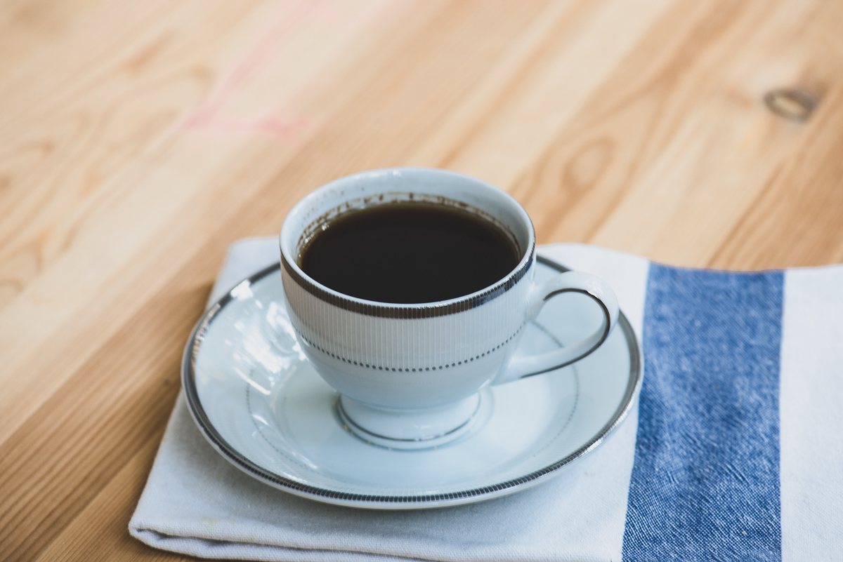 Fotografie reprezentand o ceasca de cafea plina pe o farfurioara alba. Dedesubtul acesteia se afla un servet de bucatarie alb cu albastru